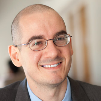 A headshot of Angelo Mele. He has a shaved head and oval glasses. He is wearing a suit and in front of a blurred-out interior background.