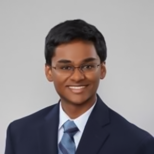 A headshot of Sampath Rapuri. He is wearing a navy suit jacket and against a plain gray background. He has short, dark brown hair, glasses, and is smiling.