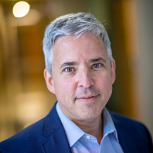 A headshot of Dr. G Caleb Alexander, who has short gray hair, light skin, a navy jacket and a blue collared shirt. The background is out of focus.