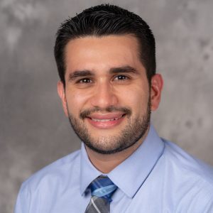 A headshot of Corey Oses who is against a plain background. He has short dark hair and is wearing a collared shirt and tie.