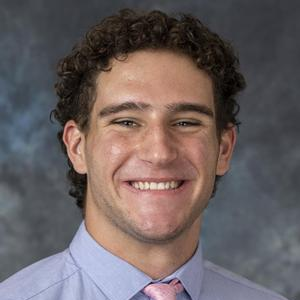 A headshot of William Shiber. William is grinning and has short curly hair. He is wearing a light blue collared shirt and pink tie and is against a blue abstract background.